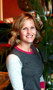 A head-and-shoulders shot of a young woman with a name tag posing in a hotel lobby.