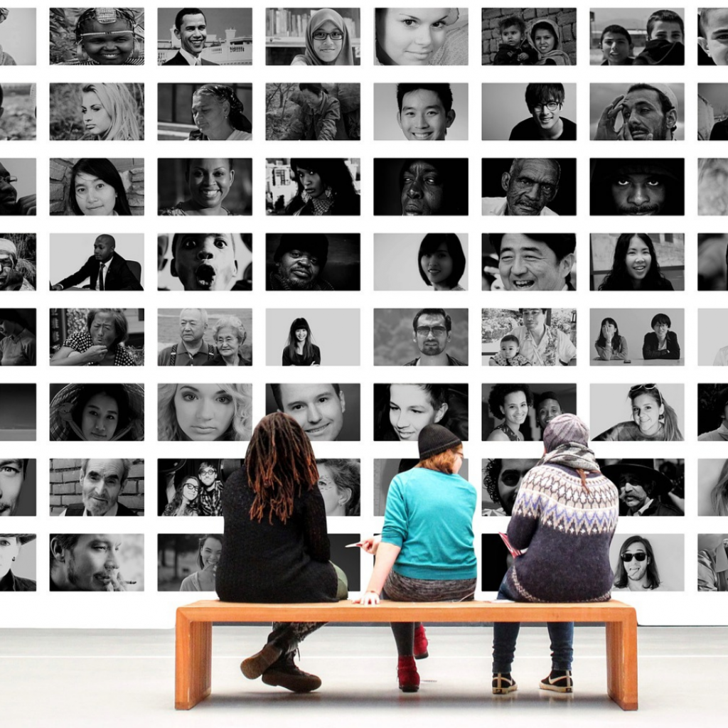 Three people sit on a bench looking at a large gallery of black and white photos of different people.