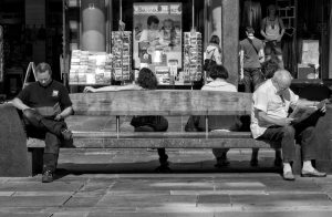 Two people at either end of a long bench, each reading a newspaper.
