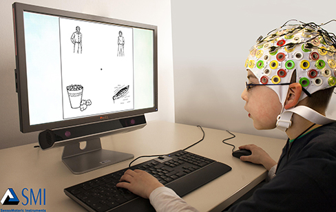 A photograph depicts a person looking at a computer screen and using the keyboard and mouse. The person wears a white cap covered in electrodes and wires.