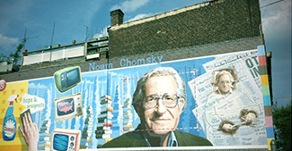 A photograph shows a mural on the side of a building. The mural includes Chomsky's face, along with some newspapers, televisions, and cleaning products. At the top of the mural, it reads “Noam Chomsky.” At the bottom of the mural, it reads “the most important intellectual alive.”