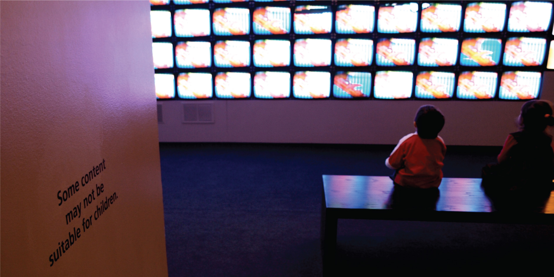 Children sit in front of a bank of television screens. A sign on the wall says, “Some content may not be suitable for children.”