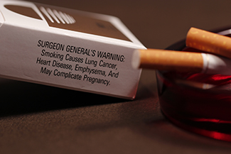 A photograph shows pack of cigarettes and cigarettes in an ashtray. The pack of cigarettes reads, “Surgeon general’s warning: smoking causes lung cancer, heart disease, emphysema, and may complicate pregnancy.”