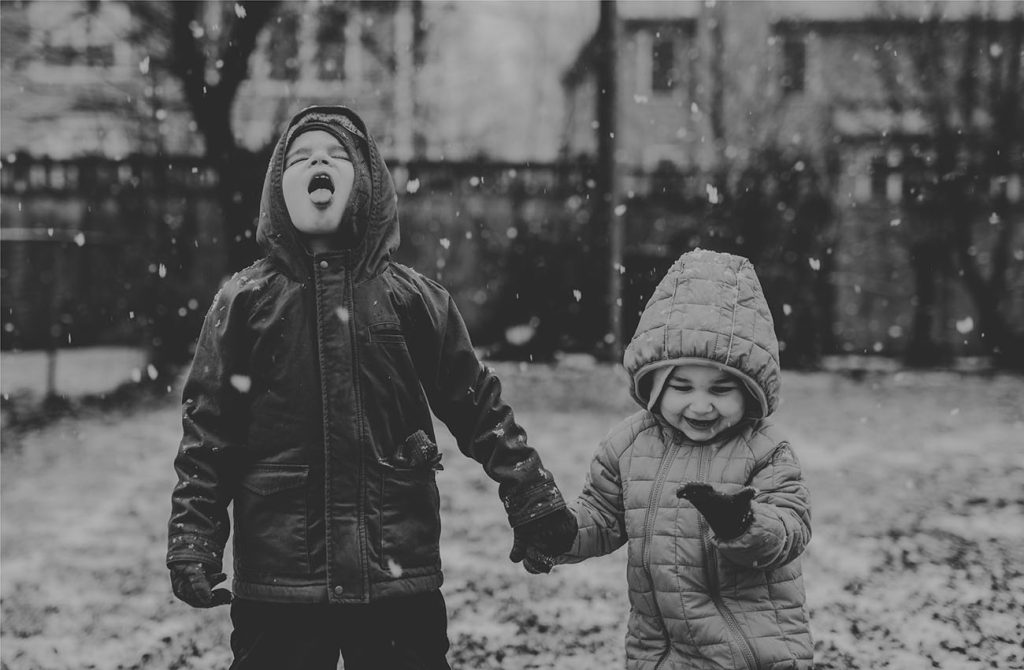 A greyscale photograph shows two children standing in falling snow. The child on the left has their mouth open, catching snowflakes on their tongue. The child on the right has their palm open and is looking at snowflakes on their mitten.
