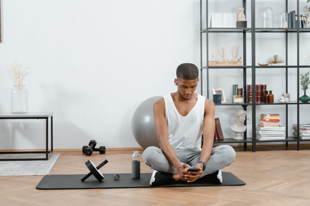 Man sitting on yoga mat with exercise equipment. The man is looking at his phone.