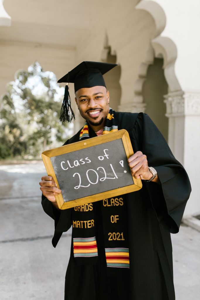 Photograph of a man in graduation gown and cap with scarf that says "Black Grads Matter" and "Class of 2021". He is holding a small chalkboard with 'Class of 2021' written on it.