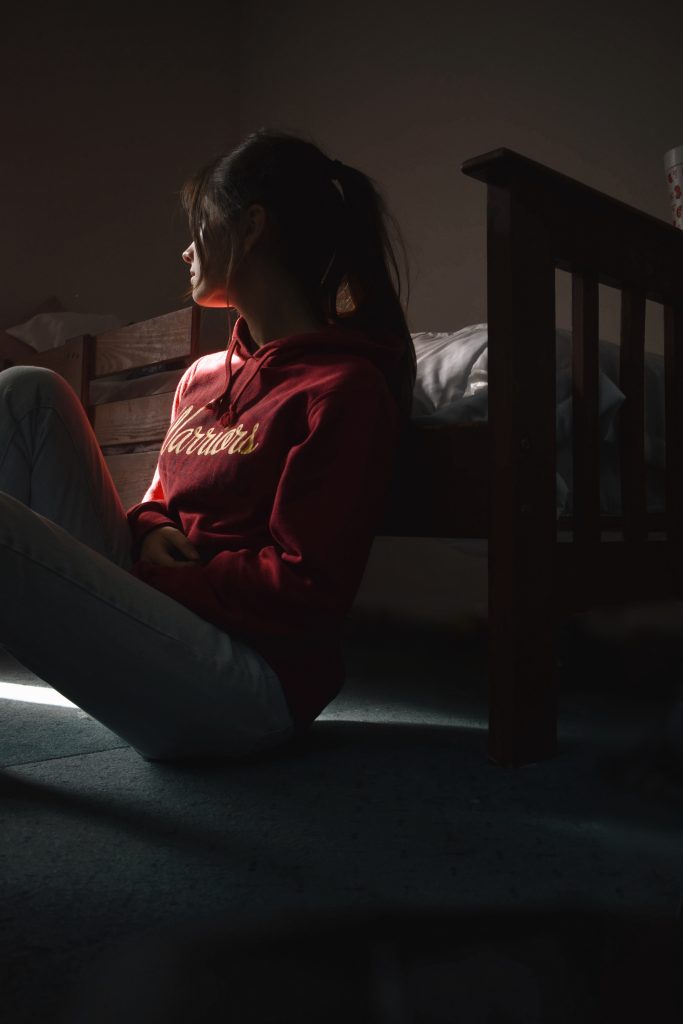 In a darkened room, a university student sits on the floor leaning against the bed and looks away from camera and towards a light source.