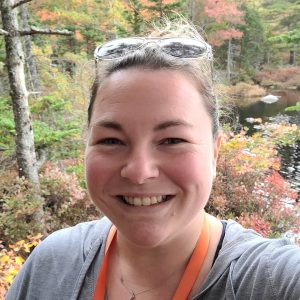 A smiling brown haired woman with a grey shirt, hair pulled back and sunglasses on her head. She is standing in the middle of a wooded area, behind her you can see a river.